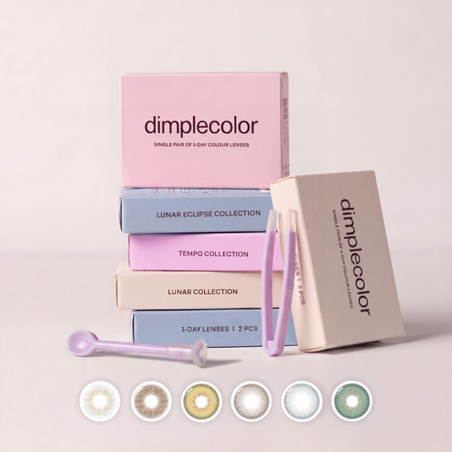 A set of Dimple Color contact lens boxes and accessories arranged on a light beige background. The stack includes pink, blue, lavender, and cream boxes with a small applicator tool and tweezers.
